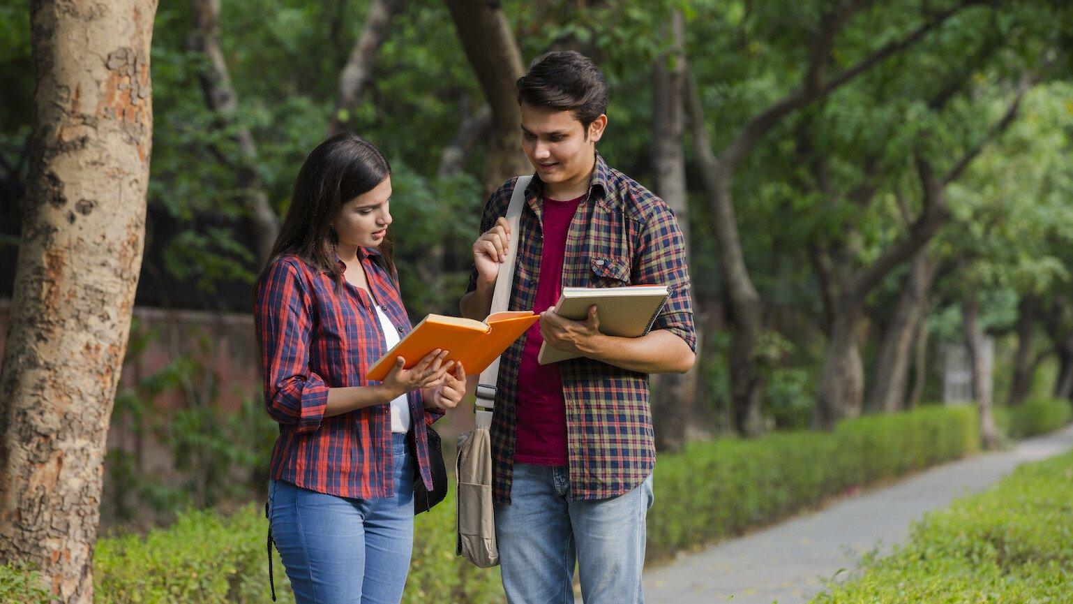 Two students talking to each other and looking at a book. They are stood outdoors on a path lined by trees.